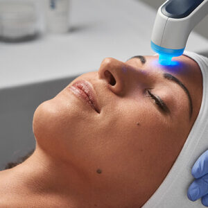A woman receiving a facial light therapy treatment with a blue light device pressed to her forehead, eyes closed, wearing a headband.